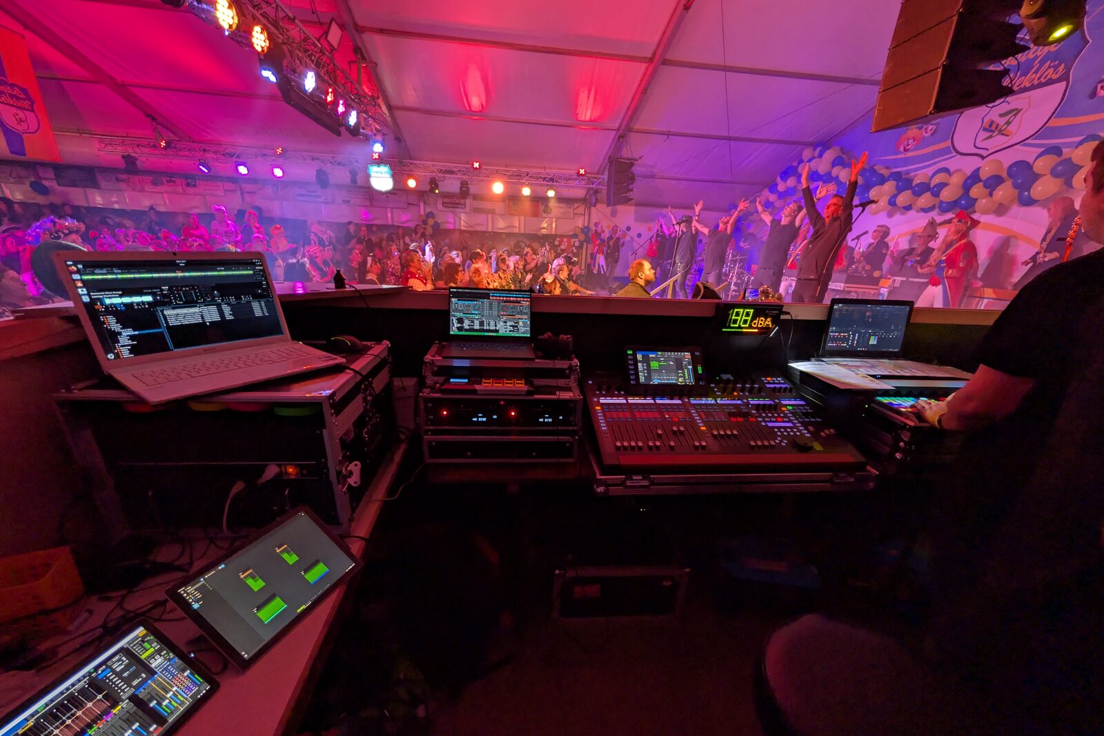 Front-of-house desk with digital mixing console, playback laptops and a level meter, band on stage in a festival tent